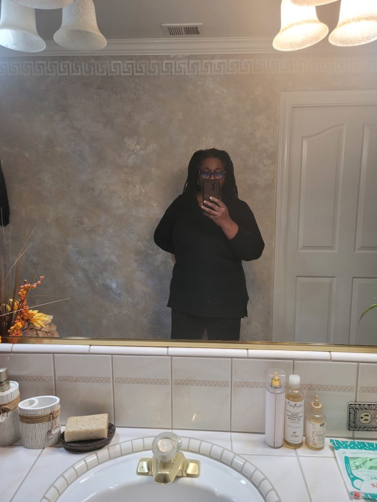 Fitness journey for week 2. Woman (myself) standing in front of mirror in a black sweater and black pants. Hair let down. Showing week 2 body size