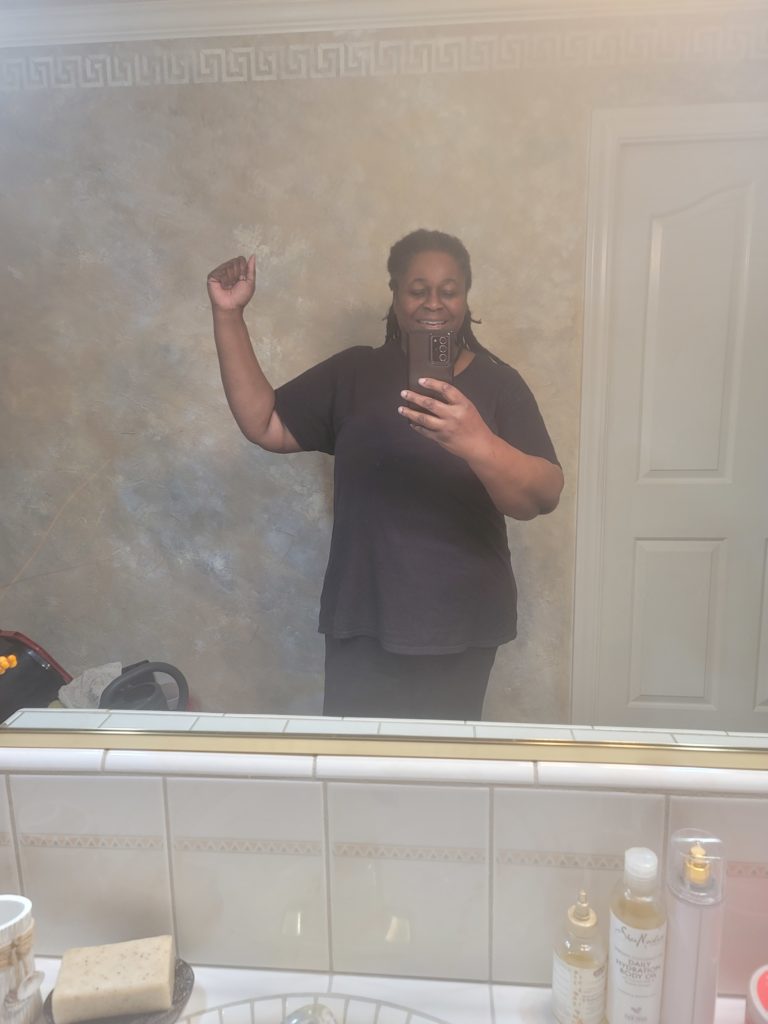 Fitness journey of woman facing mirror with right hand raised to shoe size of arm. Woman is wearing a black t-shirt and black pants.