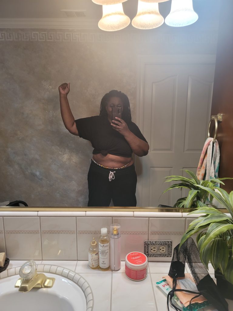 Fitness Journey of woman facing the mirror with right hand raised and mid-section revealed to show the size of waist line