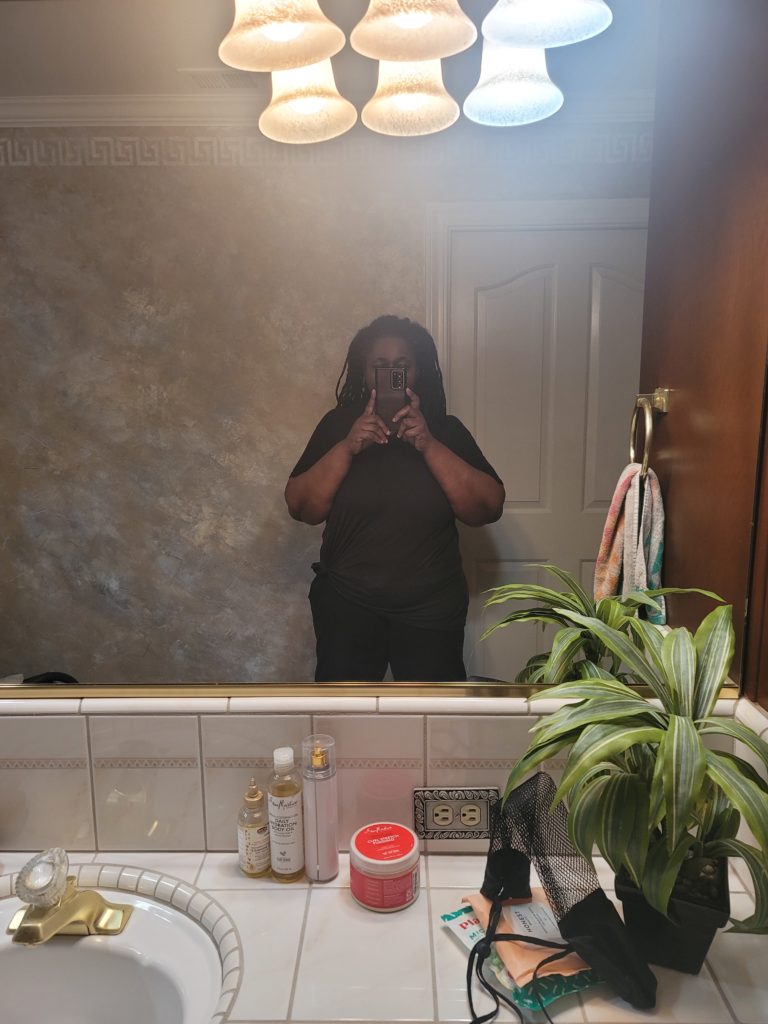 Self of current size facing a very wide bathroom mirror. Lady wearing a black T-shirt and black 